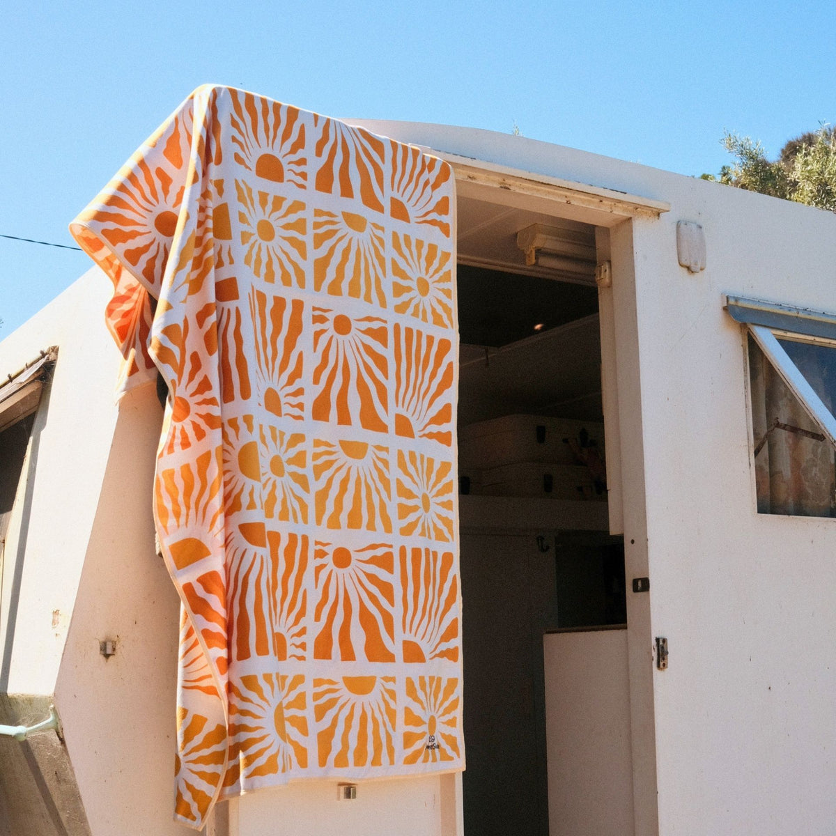Solstice sand free beach towel in Large, yellow retro sunburst print, draped on a caravan against blue sky