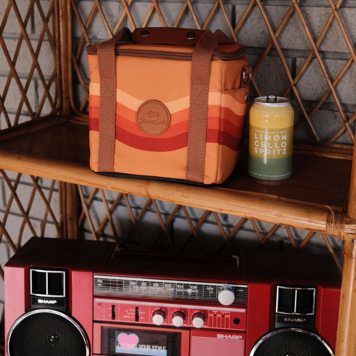 Stevie mini insulated cooler bag, orange retro 70s wave print, on a cane shelf with a drink