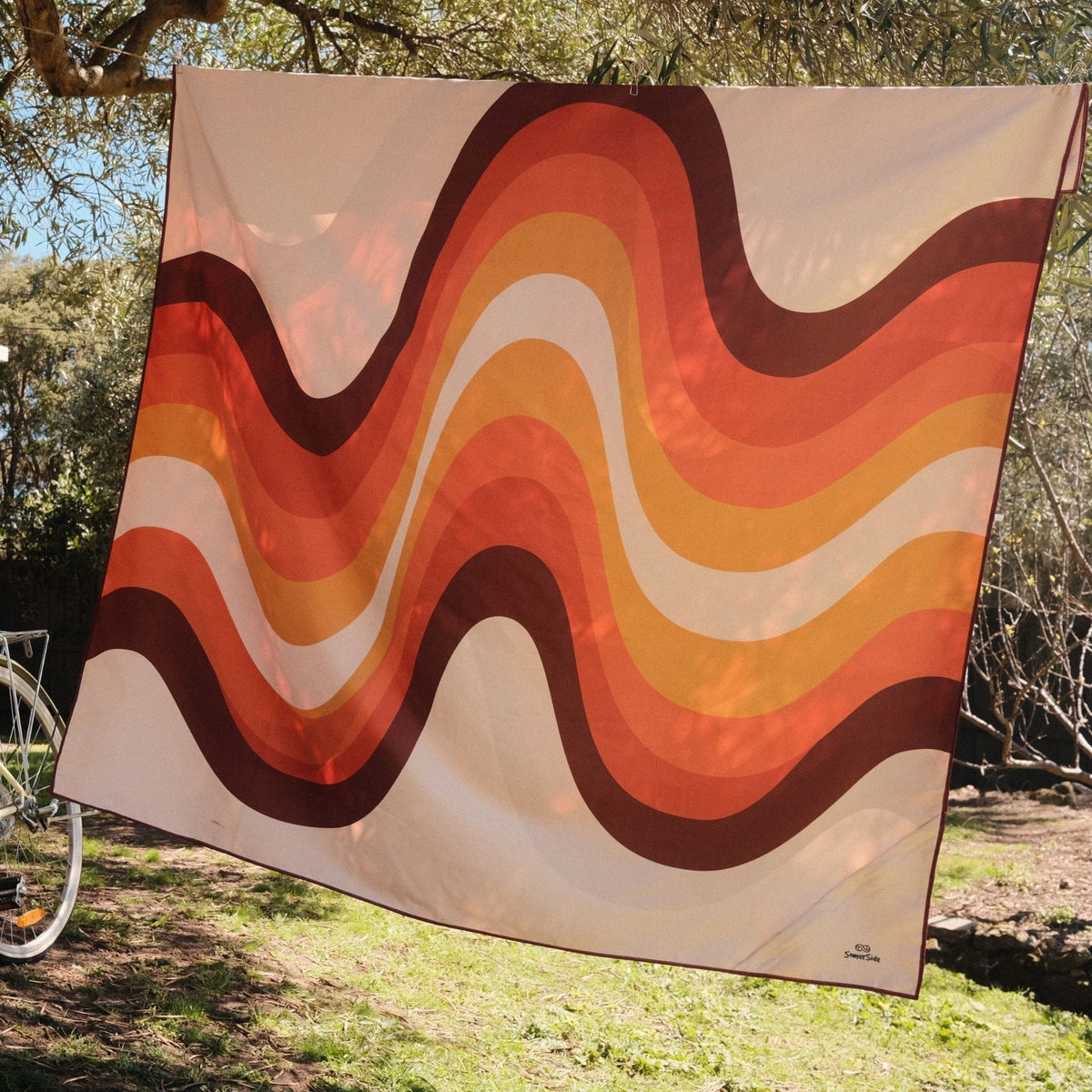 Stevie sand free beach towel in XL, orange retro 70s wave print, on a washing line in the sun