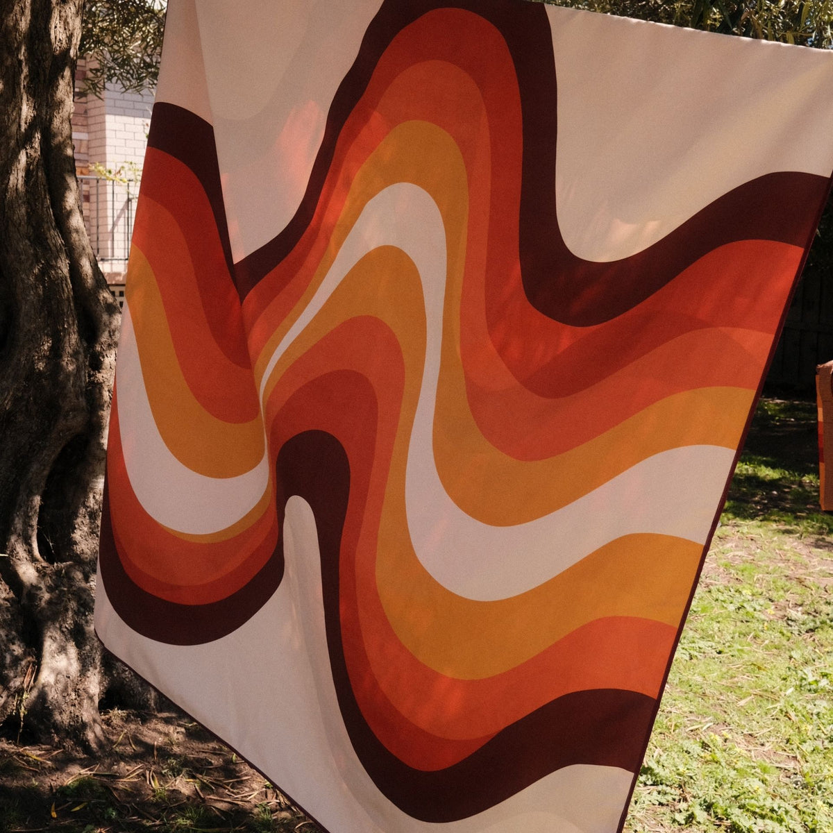 Stevie sand free beach towel in XL, orange retro 70s wave print, on a washing line under a tree