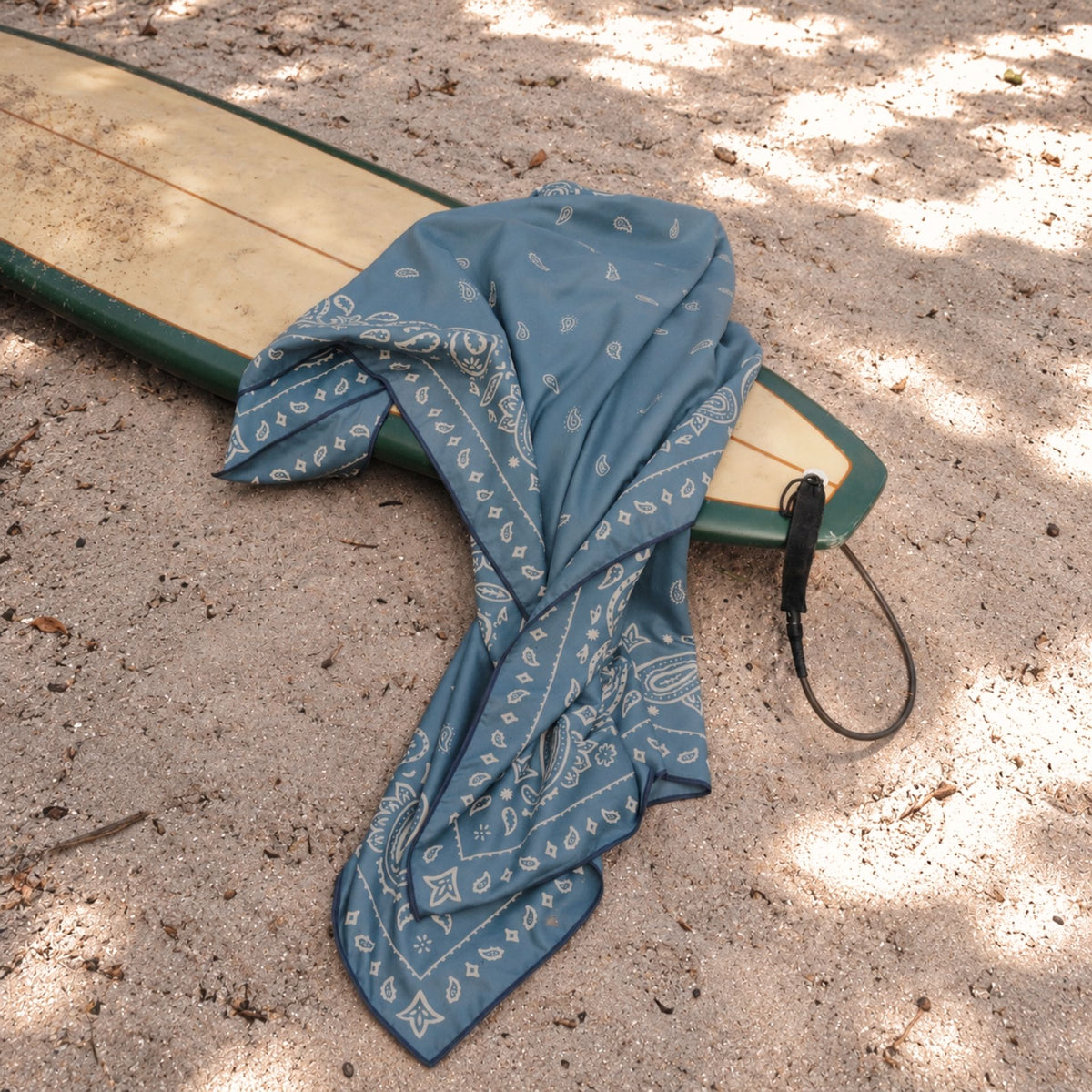 Venice sand free beach towel, dusty blue paisley bandana print, on a surfboard in the sand