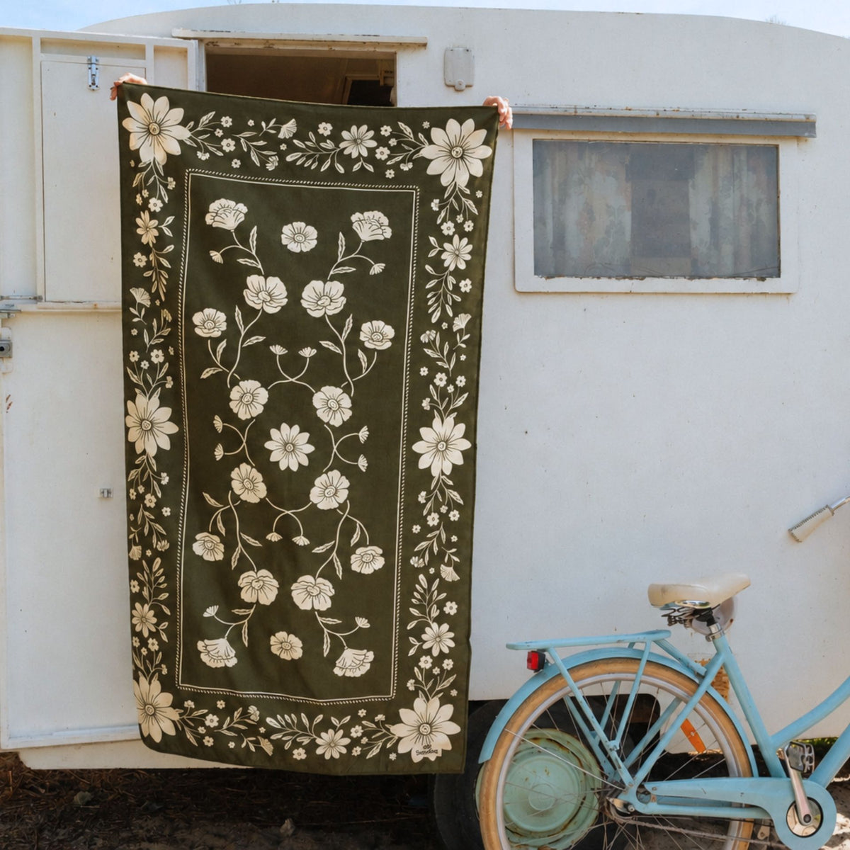 Eden sand free beach towel in Large, olive green floral bandana print, hung on a caravan by a bike