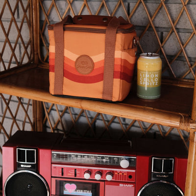 Stevie mini insulated cooler bag, orange retro 70s wave print, on a cane shelf with a drink