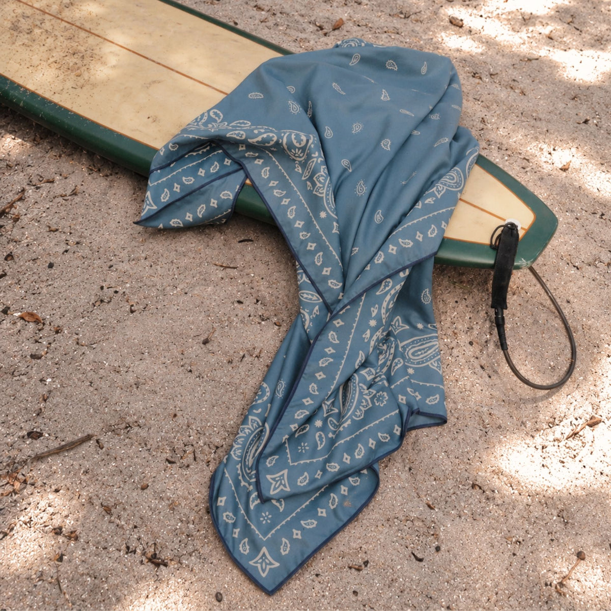 Venice sand free beach towel, dusty blue paisley bandana print, on a surfboard at the beach