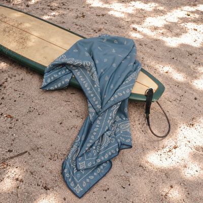 Venice sand free beach towel, dusty blue paisley bandana print, on a surfboard in the sand